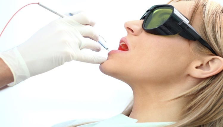 A blonde woman in green safety glasses gets a laser treatment on her mouth from a gloved hand holding a device